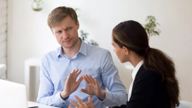 Man and woman sitting at desk in office arguing, man with hands up in frustration.