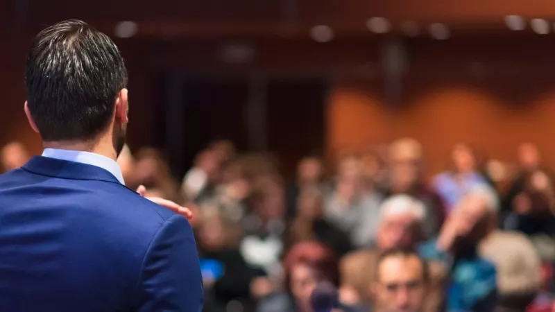 A man stands before a crowd of people in a room, with a blurred background behind him.