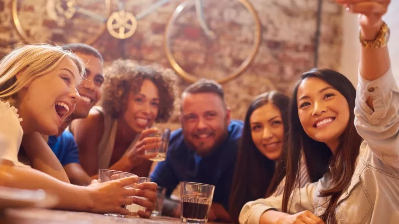 Group of friends smiling and taking a selfie while sitting at a table in a bar.