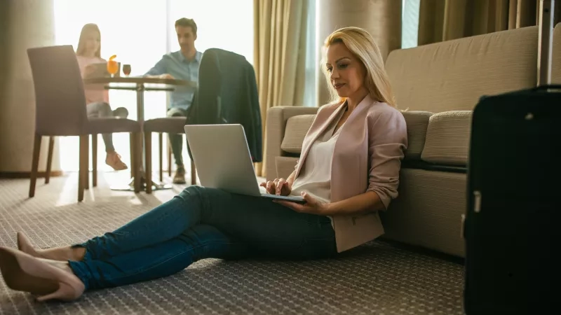 Woman on laptop in a hotel room, with a couple dining in the background.
