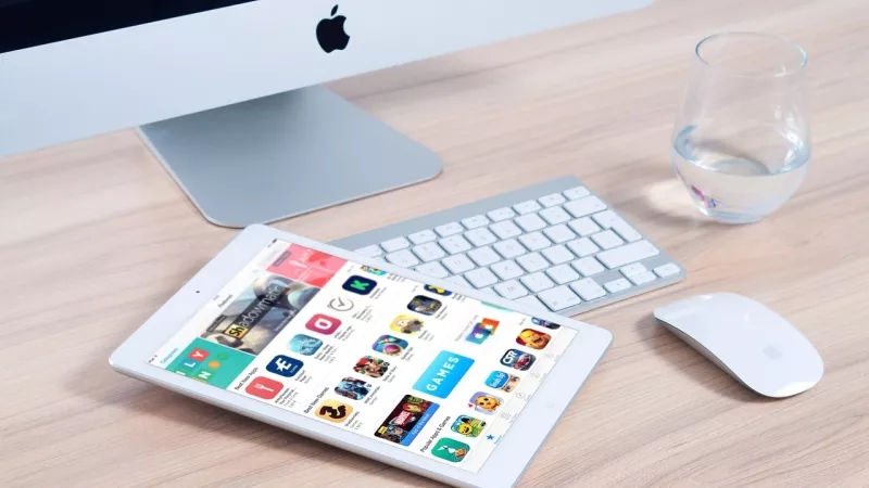 Apple iPad and Apple iMac on a table with a glass of water and an Apple wireless mouse.