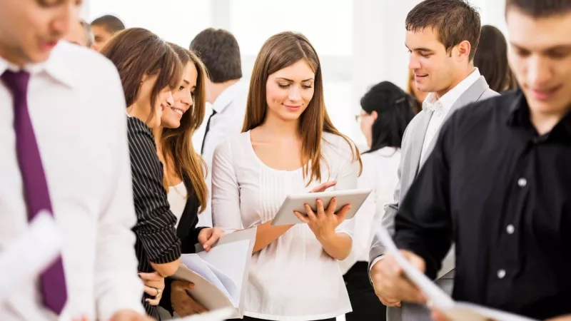 Woman looking at her tablet while people are reading papers in front of her in a business meeting.