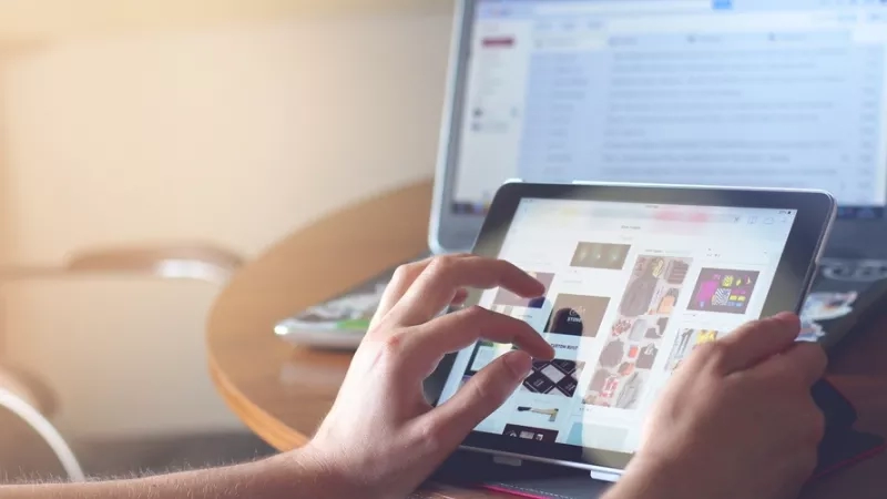 A person using a tablet device to touch and scroll through the screen with a laptop in the background.