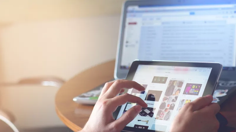 A person is using a tablet to shop online at a table with a laptop open in the background.