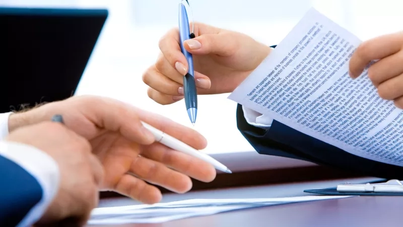 Business people sign contracts in a meeting room, holding pens, and a laptop in the background.