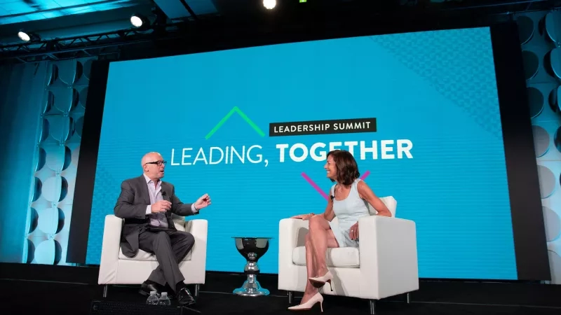 Two people sit on chairs on a stage in front of a large screen saying Leadership Summit.