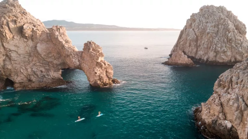 Two people on surfboards in the blue ocean with rock formations on the side.