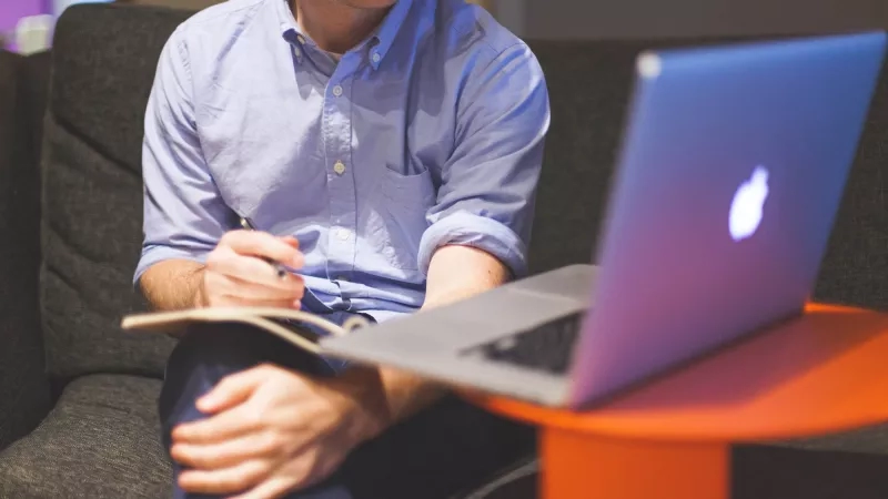 Man sitting on couch with pen and paper and laptop on orange side table next to him.