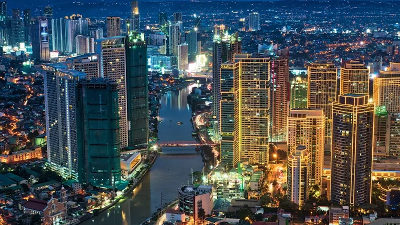 A vibrant cityscape of Manila, Philippines, with towering buildings and a winding river at night.