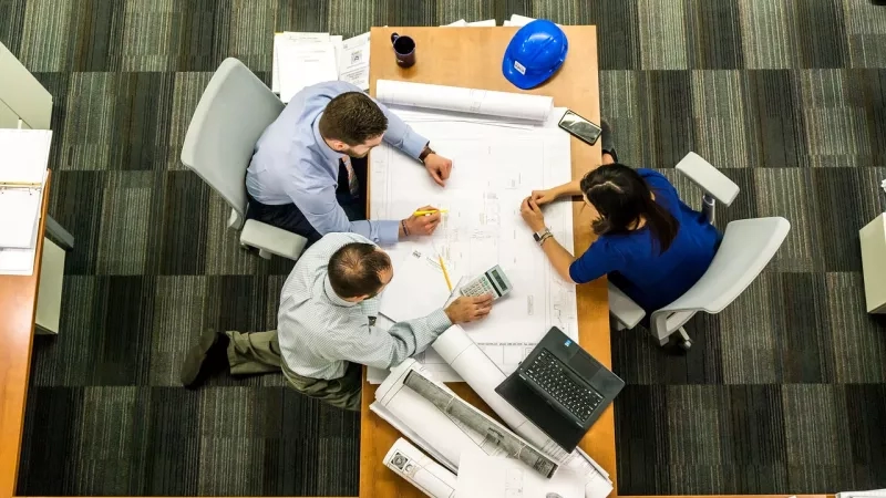 Overhead view of a meeting with three people working on blueprints, a laptop, and other items on a table.