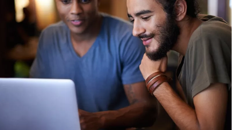 Two men looking at a laptop with a piece of paper on a table in front of them.