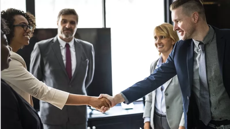 A group of business people shake hands in an office with a large monitor in the background.