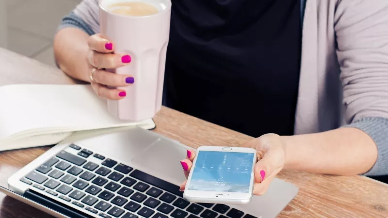 Freelancer with pink nails holding coffee and smartphone in office with laptop and open book on desk.