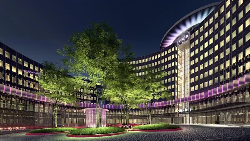 Park Inn Amsterdam building in the evening with purple lighting, trees, and a circular fountain in the foreground.