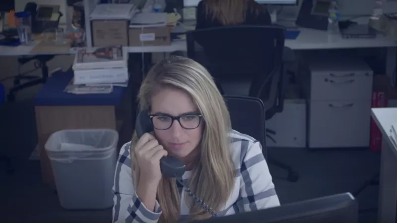 Woman with glasses talking on the phone in the office next to a trash can.