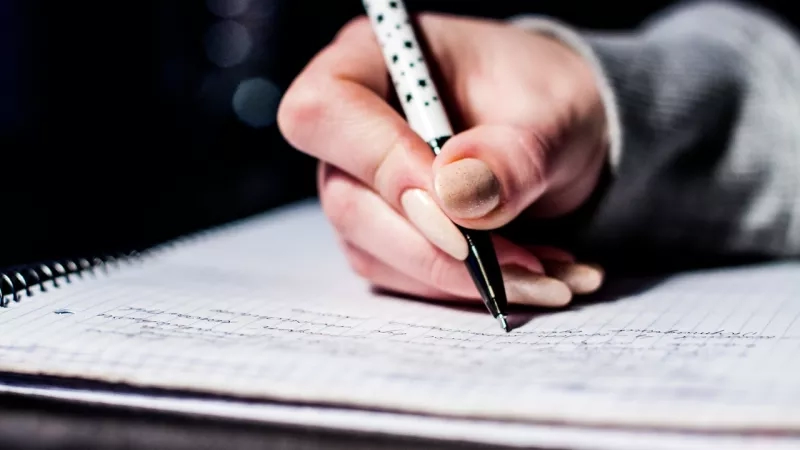 A hand holding a pen is writing notes on a spiral notebook.