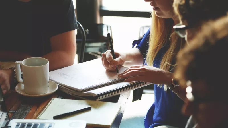 A woman is writing in a notebook while sitting at a table with others during a meeting.