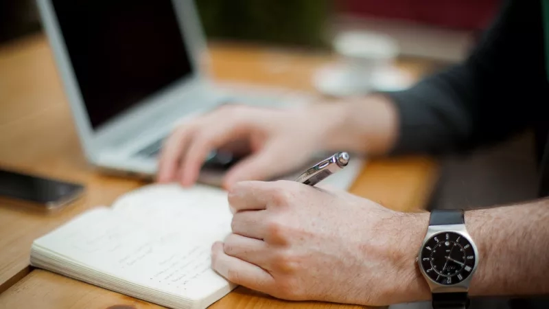 Person typing on Apple laptop and writing in notebook with pen.