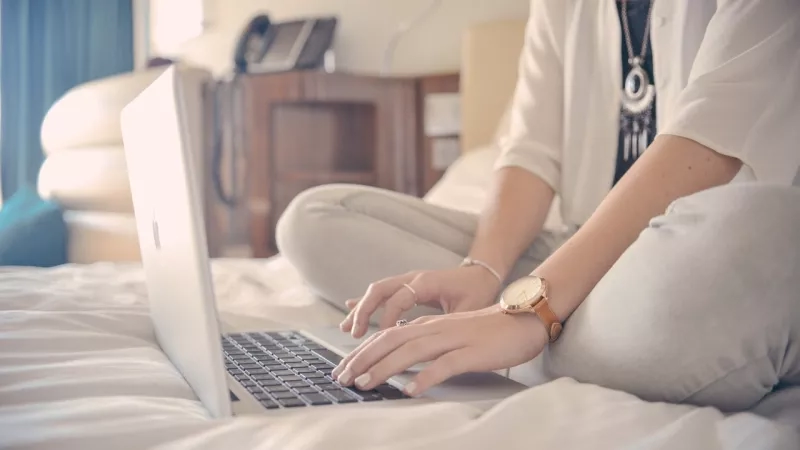 A woman is sitting on a bed using an Apple laptop in a hotel room.