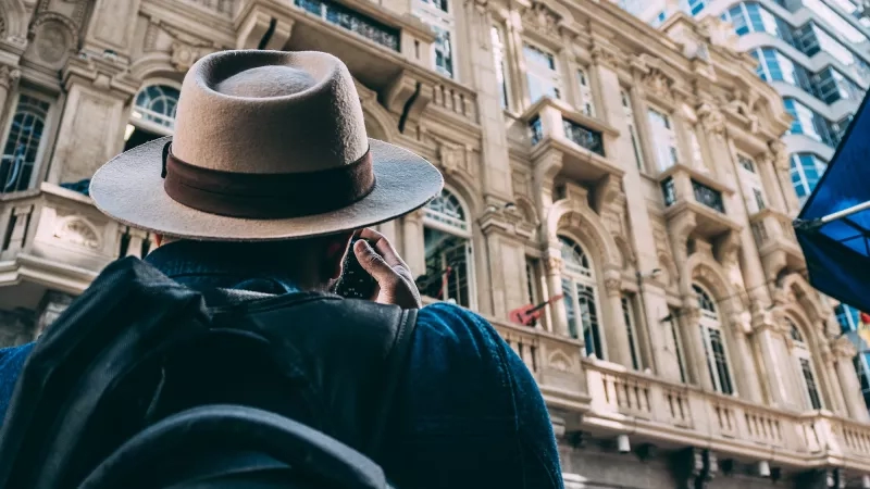 A man with a hat and backpack is talking on his cellphone in front of an architectural building.