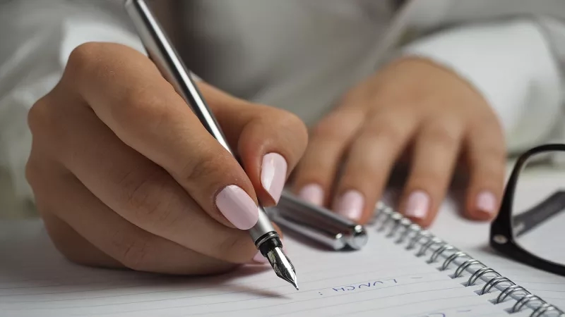 A woman writing with a fountain pen on a lined notebook with the words "Client Survey" written on it.