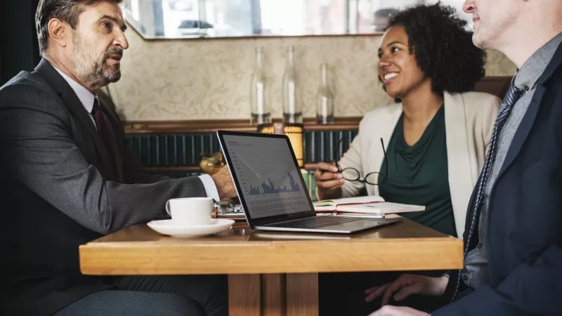 Three business people sitting around a table with a laptop showing a graph on the screen.
