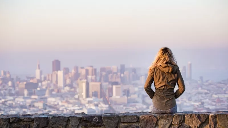 Woman sitting on a stone wall overlooking a city with tall buildings and a hazy sky at sunset.