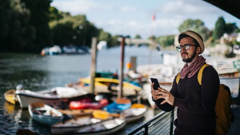 A man with a hat is standing on the pier near boats while looking at his cellphone.