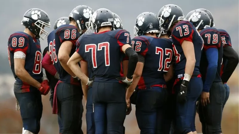 American football players wearing uniform and helmet gathered in circle, perhaps during a huddle on the field.