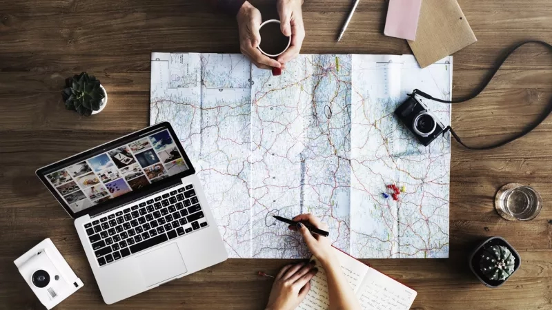Overhead view of people planning a trip with map, laptop, and journal on wooden table.