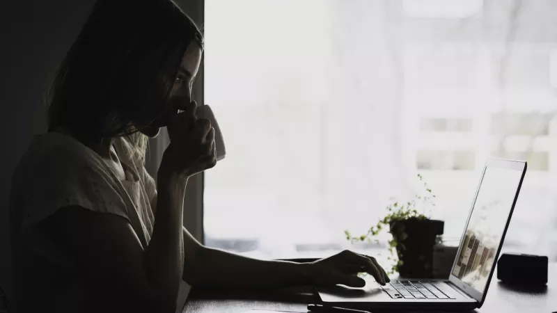 A woman using her laptop in a dark room with a cup in her hand.