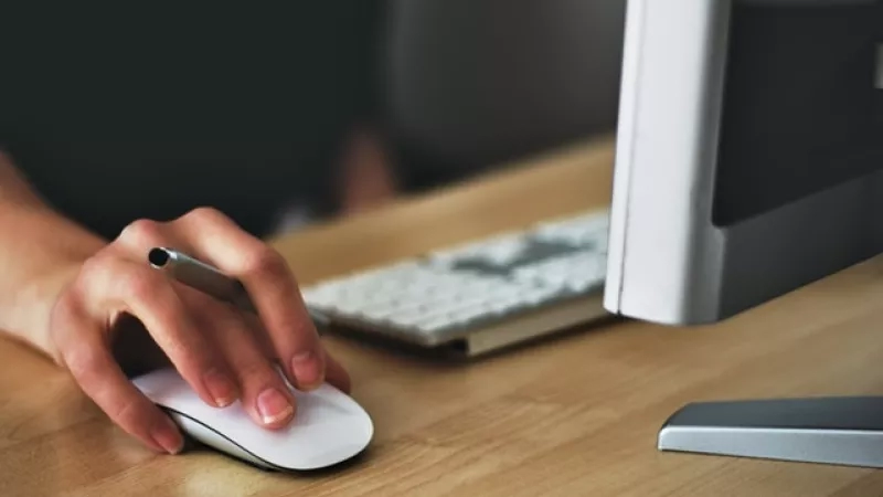 Person's hand using a mouse while sitting in front of a computer in a blurred background.