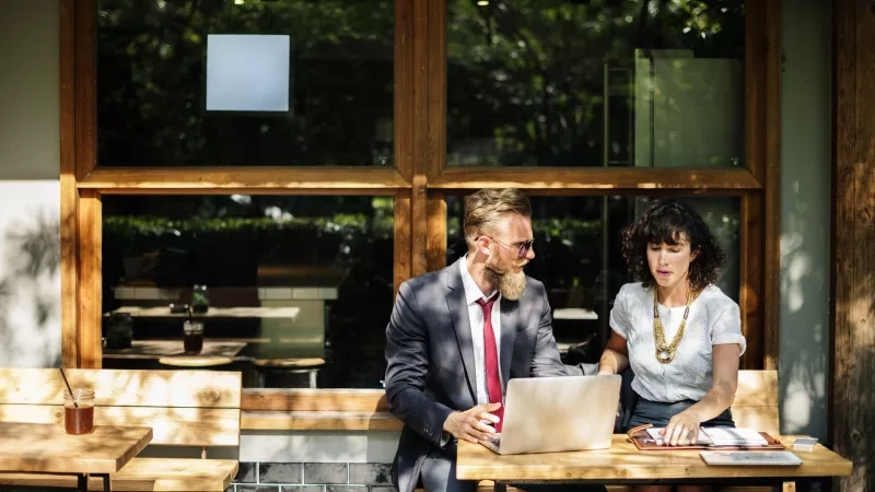 Two people are working on laptops at a table in an outdoor cafe with glass windows.