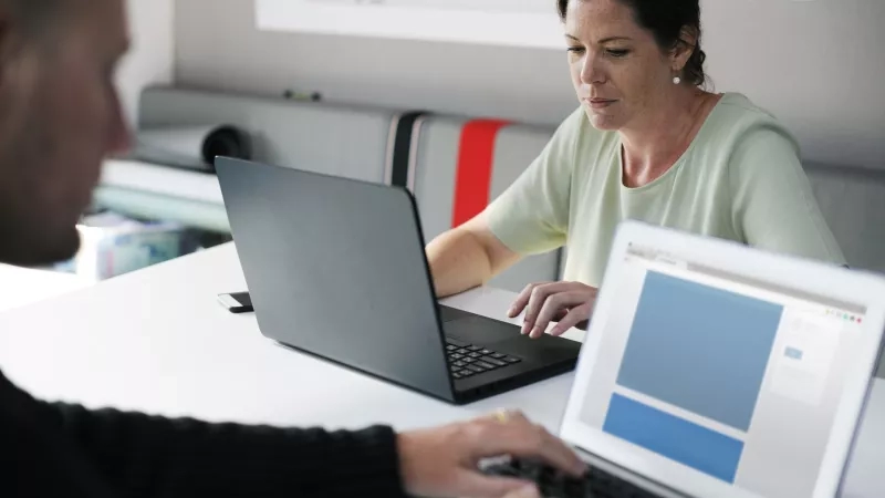 Two people are working on their laptops in a shared office space with gray walls and furniture.