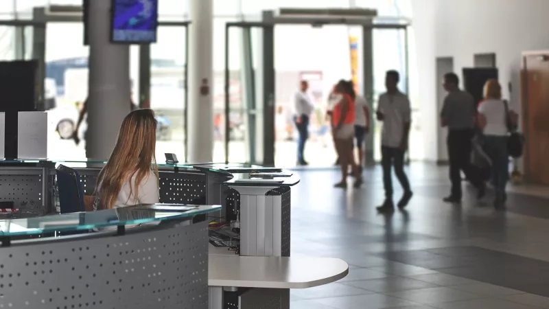 A woman at a reception desk with blurred people in the background in a public area.