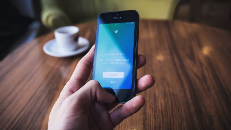 A person is holding a smartphone with the Twitter app on a table in a cafe.