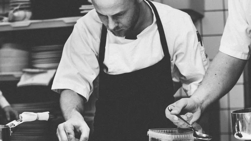 Chef in kitchen with apron on, holding knife and cutting food, with other chef beside him with spoon.