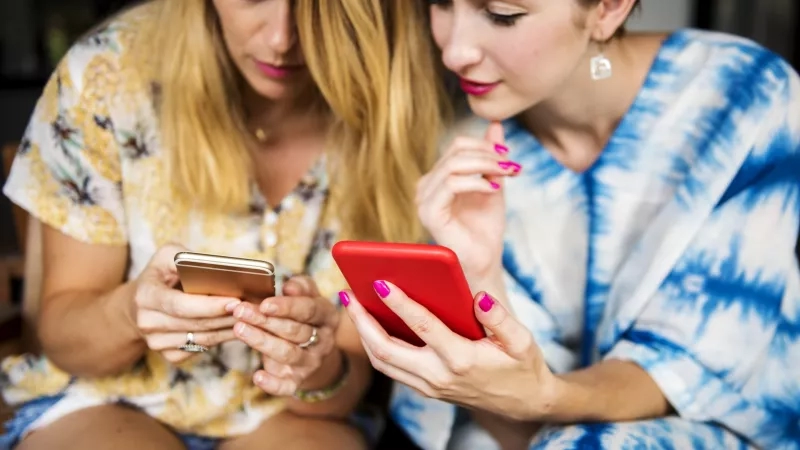 Two women in casual dress are looking at their phones with a red and a gold phone.