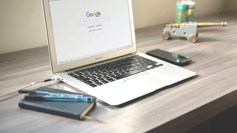 MacBook Air on a desk with a notebook, pens, cell phone, and toy cannon with a bottle.