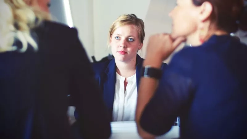 Three women are sitting in a conference room and are talking to each other about their work.