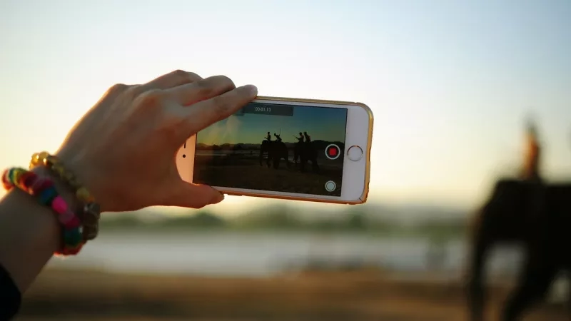 A hand holds a smartphone recording a video of people riding elephants on a sunny day.