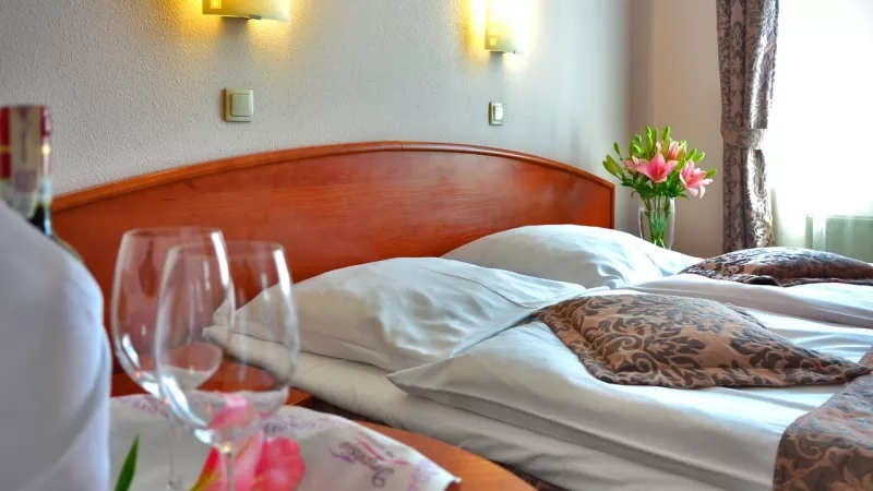 A hotel room with a bottle of wine, two wine glasses, and a bed with white linens and flowers.