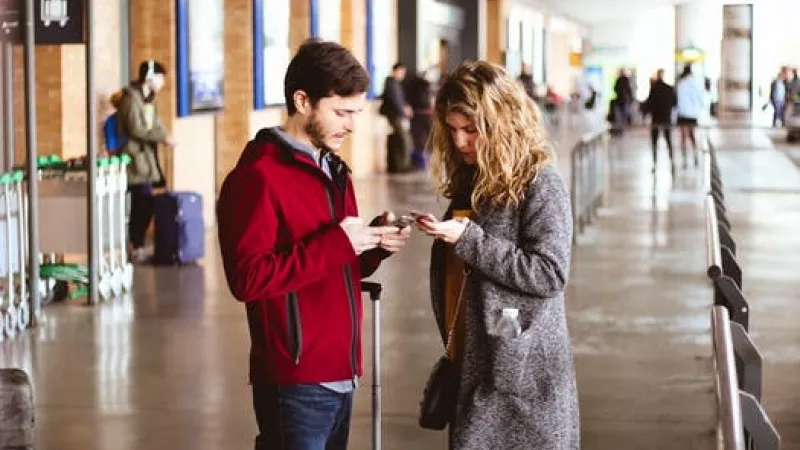 Couple standing in airport while looking at their phones with luggage behind them and others walking around.