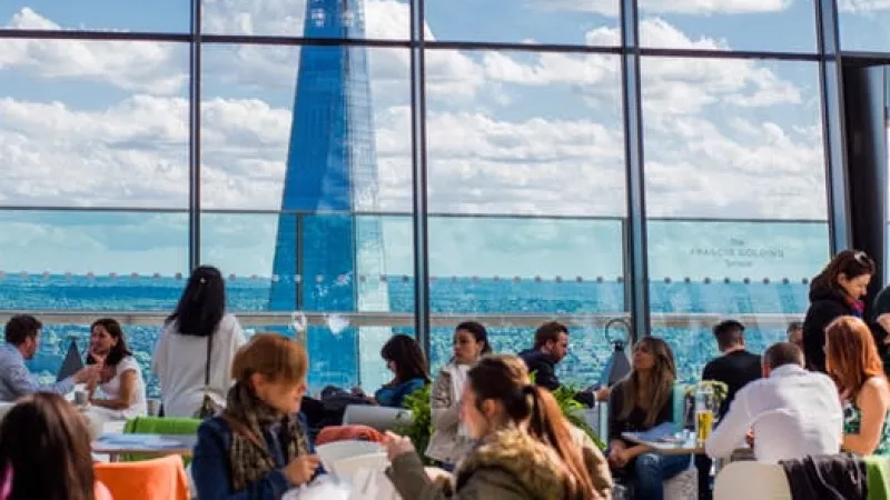 Several people sitting at tables at the Shard with the view of the Shard in the window.