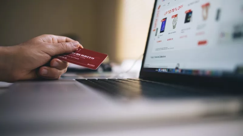 Person holding a credit card in front of a laptop displaying an online shopping site.