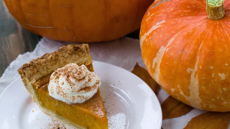 Pumpkin pie with whipped cream on a plate with autumn leaves and pumpkins in the background.