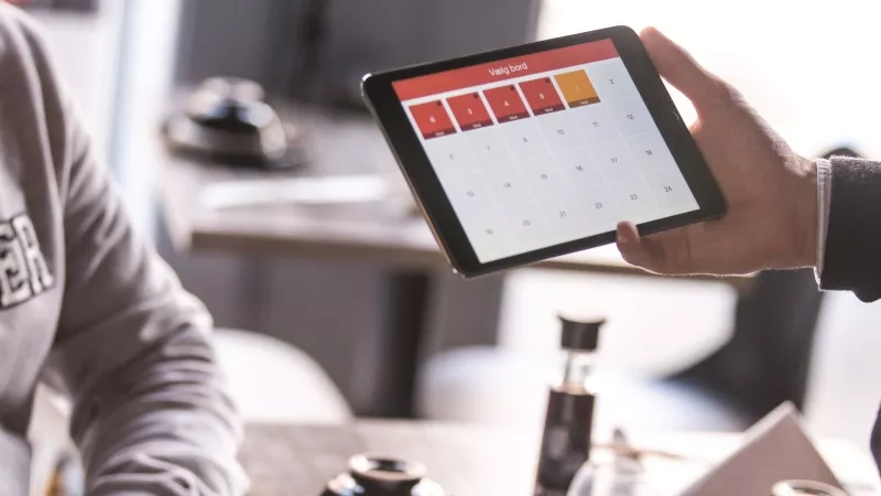 Person holds a tablet with a calendar with colored days in front of another person.