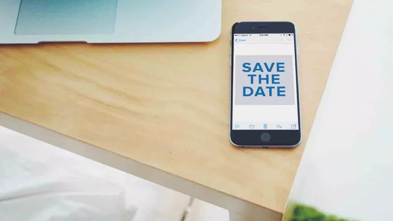 Save the date text on an iPhone, placed on a table with a laptop and white background.