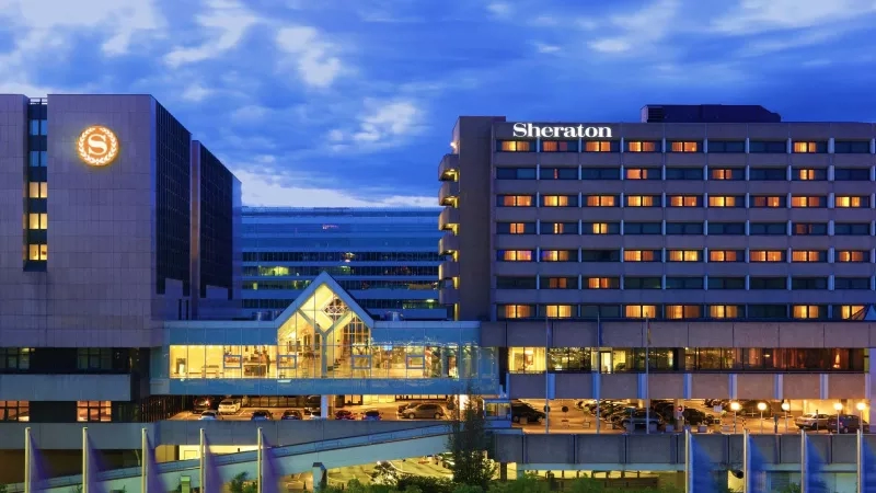 Exterior of Sheraton Frankfurt Airport Hotel with blue sky in the background and cars parked in front.