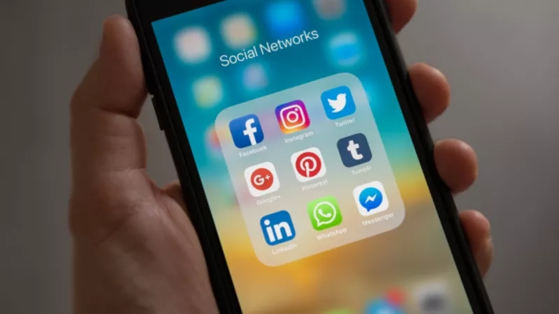A person's hand holds a smartphone displaying various social media icons.